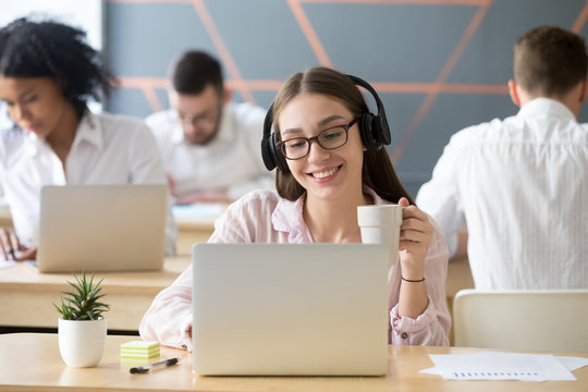 Millennial smiling woman wearing headphones watching online funny video looking on laptop in coworking at coffee break, happy student employee in headset making videocall using computer app in office