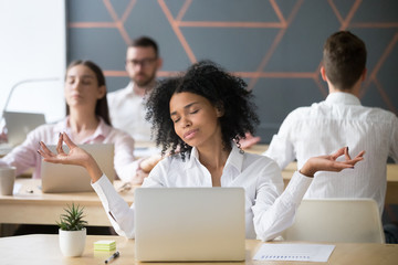 Mindful millennial african woman meditating at workplace with colleagues, calm black employee or student doing yoga exercises feeling zen enjoying no stress free relief, corporate meditation concept