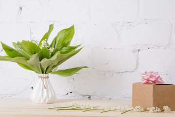 Bouquet Fresh spring lily of the valley in a white vase, Corton box with a gift and a pink bow, on a table on a white brick background. space for text