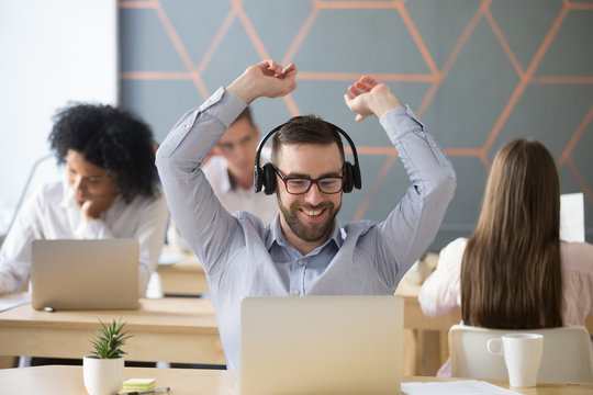 Happy businessman in headphones feeling joy celebrating success looking at laptop in coworking office, millennial manager in headset excited by online win, great deal raising hands enjoys good result