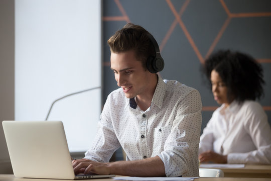 Millennial smiling man in headphones using computer software for work, study or web communication, young employee consulting client online, student in headset typing on laptop in coworking office