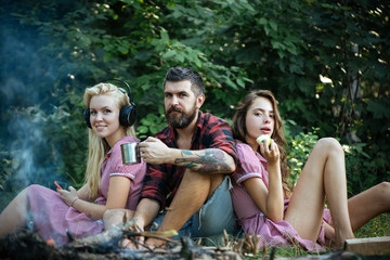 Group of friends sitting next to campfire. Girls leaning on their male friend. Blond woman listening to music while brunette lady eats apple. Smiling guy drinking tea or coffee from mug