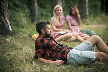 Bearded guy lying in grass on forest meadow. Girls sitting next to campfire. Friends camping in late summer. Relaxation in forest