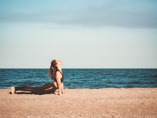 Young beautiful sporty woman in green clothing doing yoga asana on sea sandy beach near water. Girl practicing exercises. Health concept. Copy space.