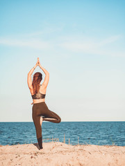 Fototapeta na wymiar Young beautiful sporty woman in green clothing doing yoga asana on sea sandy beach near water. Girl practicing exercises. Health concept. Copy space.