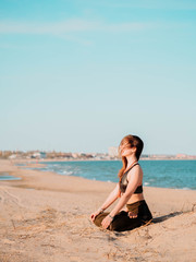 Fototapeta na wymiar Young beautiful sporty woman doing yoga asana on sea beach near water. Girl practicing meditation. City on background. Health concept. Copy space.