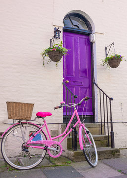 Bright Pink Woman  Bicycle With Rear Wicker Basket Parked In Front Of A Violet Wooden Front Door Of A Classic Victorian British Style House