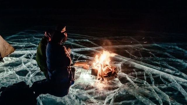 Three travelers by the fire right on the ice at night. Campground on ice. The tent stands next to the fire. People are warming themselves by the fire. Time-lapse with a circular motion. The Lake