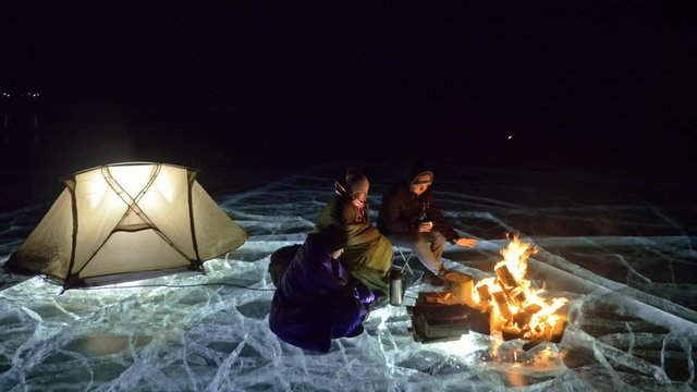 Three travelers by fire right on ice at night. Campground on ice. Tent stands next to fire. Lake Baikal. Nearby there is car. People are warming around campfire and are dressed in sleeping bags. This
