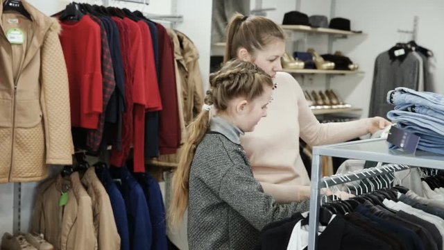 Cheerful daughter with young mother choosing new stylish clothes in retail store
