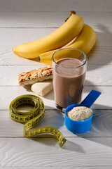 glass of protein shake with bananas and energy bars on white wooden table