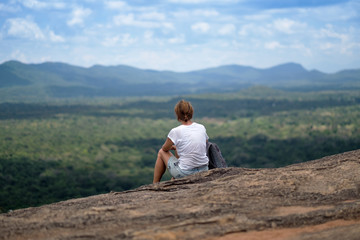 Naklejka na ściany i meble Young woman with backpack sitting on mountain and looking to a sky with clouds. Beautiful nature of Sri Lanka