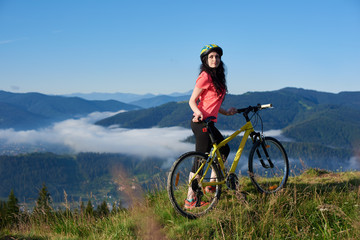 Fototapeta na wymiar Beautiful female biker with yellow bicycle in the mountains in the morning, wearing helmet and red red t-shirt. Foggy mountains, forests on the blurred background. Outdoor sport activity