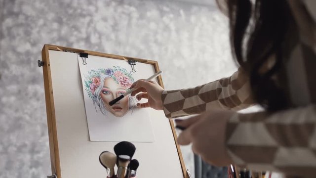 The artist on makeup and beauty creates a beautiful face makeup chart using watercolor paper, shadows, lipsticks and blushes using an easel at studio. Young woman in a bright studio draws a face chart