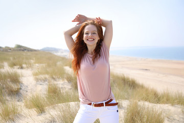 Hübsche rothaarige Frau steht in einer Dünenlandschaft und lacht Strand und Meer im Hintergrund