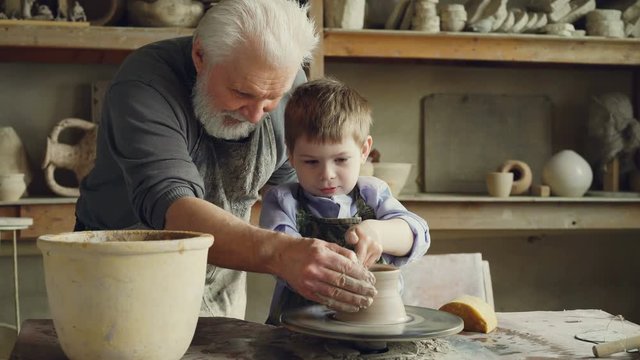 Concentrated little boy is learning to work with clay on throwing-wheel in pottery class in traditional workshop. His teacher senior experienced man is helping him.