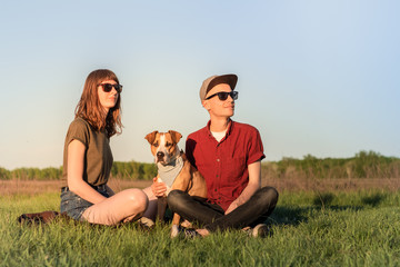 Young happy couple of man and woman with staffordshire terrier dog sit at lawn. Two persons in casual clothes hug adopted pitbull dog in bandana and enjoy beautiful sunny afternoon in spring or summer