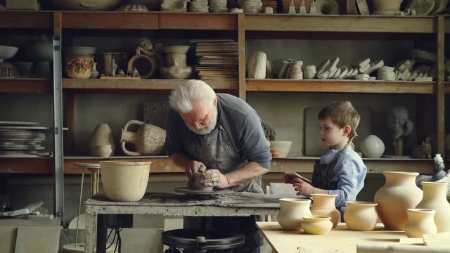 Professional male potter is working with clay on spinning throwing-wheel with his curious grandson helping him. Ceramic pots, vases and figures are visible.