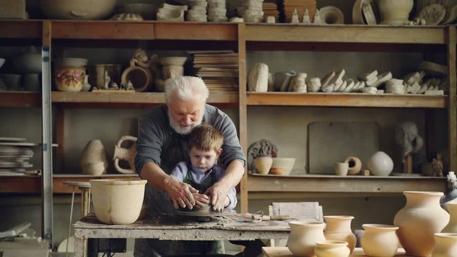Loving grandfather professional potter is teaching his small grandchild to mold clay on potter's wheel. Little boy is laughing and enjoying new hobby.
