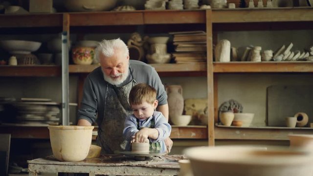 Elderly man experienced potter is teaching little boy how to work with clay on potter's wheel. Sharing experience, family tradition and modern pottery concept.
