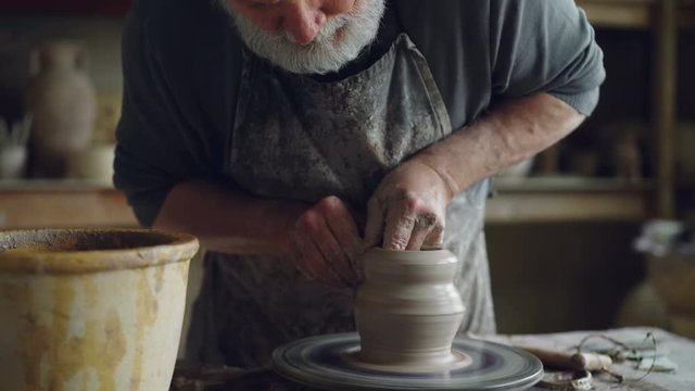 Tilt-up shot of professional potter creating jar from brown clay in workplace using throwing-wheel. Traditional pottery, handmade utensils and hardworking people concept.