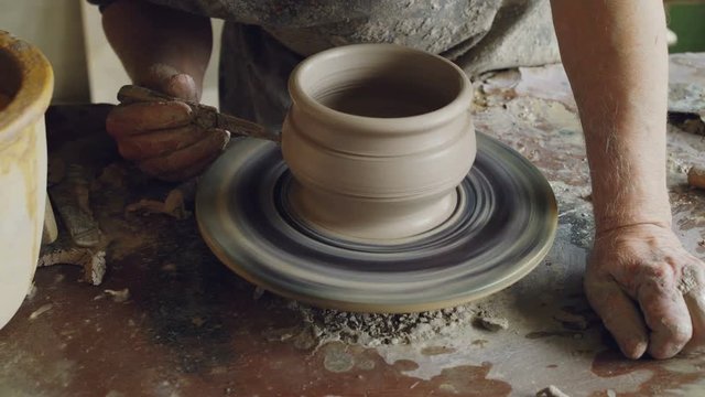Close-up shot of male hands decorating ceramic pot with knife on spinning potter's throwing wheel, making ornament. Pottery, hobby and handicraft concept.