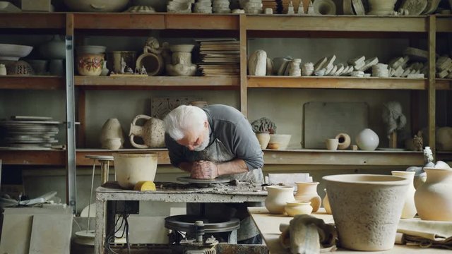 Hardworking silver-haired man is working with clay on potter's wheel, shaping piece of loam. Beautiful ceramic utensils, handmade pots and vases on shelves are visible.