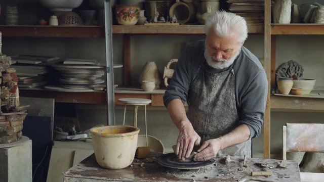Creative male potter is forming unsymmetrical broad bowl on throwing wheel while working in small workshop. Creativity, pottery and unusual hobby concept.