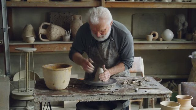 Creative senior potter is wetting hands in bowl with water and touching piece of clay on spinning throwing-wheel. Bearded man is concentrated on work.