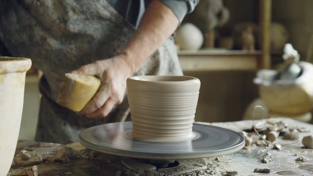 Close-up of male hands decorating half-finished ceramic pot on spinning potter's throwing wheel, making ornament. Pottery, hobby and handicraft concept.