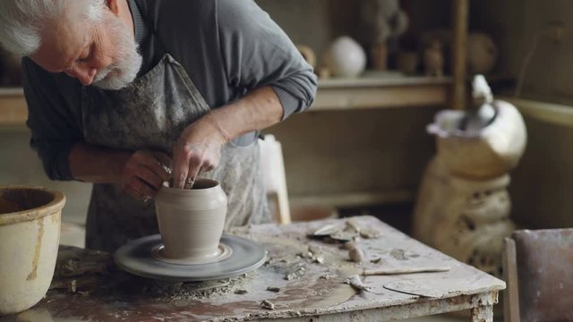Experienced ceramist grey-haired bearded man is smoothing molded ceramic pot with wet sponge. Spinning throwing wheel, muddy work table and clayware are visible.