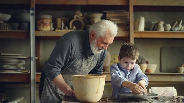 Pottery teacher senior adult is helping young student to form pot from piece of clay on throwing-wheel. Cute boy is awkward, and craftsman is helping him.