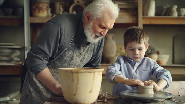 Concentrated young boy is molding clay into ceramic pot on spinning throwing wheel and his experienced grandfather is talking to him. Pottery and family tradition concept.