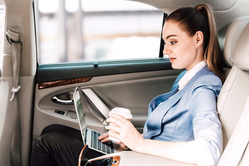 Businesswoman work with laptop computer and holding cup of coffee in back seat of luxury car