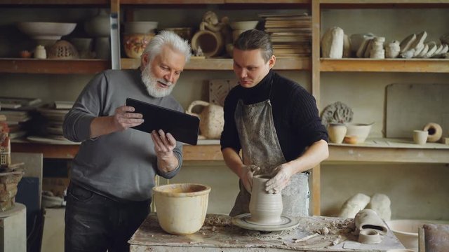 Young male sculptor is making clay figure on throwing wheel while his father is showing him tablet, men are discussing pottery and watching screen together.