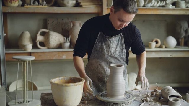 Handsome young man is molding ceramic vase from clay on spinning throwing wheel while working in potter's studio. Professional is concentrated on work.
