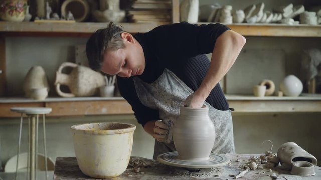 Skillful young potter is molding ceramic vase from clay on turning throwing wheel while working in potter's workshop. Professional is concentrated on work.