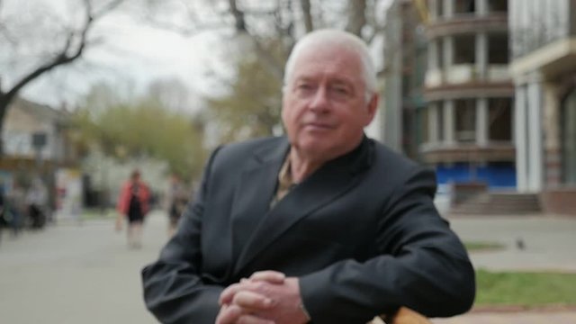 A zoom in shot of a confident white-headed man sitting on a bench in a city street with bare trees and modern houses in spring in slo-mo 