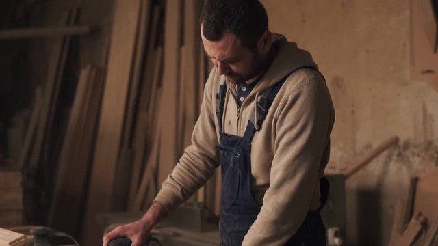 A footage of a carpenter working in overalls. A man grinding a wood in a carpentry shop. Slow motion
