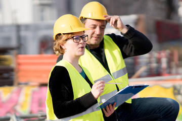 Civil engineers, a man and a woman, wearing safety jackets and helmets, checking projects on construction site