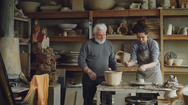 Caring senior grandfather is showing young granddoughter how to work with clay on throwing-wheel in workshop. Pottery, family hobby and handicraft concept