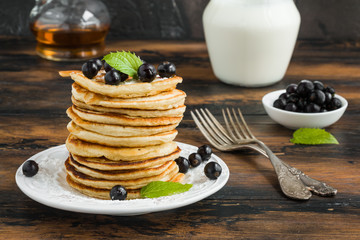 Sweet pancakes with black currant and powdered sugar on white plate, fresh berries, mint and jar of milk on wooden rustic table