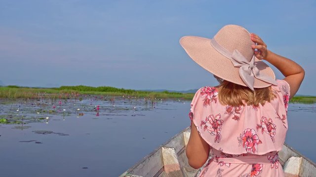 Female Tourist in Hat Riding Boat Among Red Lotus Flowers. Young Woman Visiting Thale Noi Waterfowl Reserve Lake, Thailand