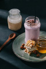 closeup view of berry smoothie with drinking straw, slices of energy bar, jam and walnuts on plate, spoon and bottle of yogurt on table