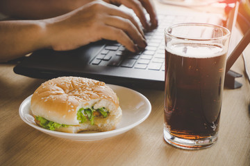business woman eating junk food burger and cold drink while working on computer laptop at office desk, unhealthy lifestyle concept