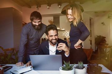Group of three young coworkers discussing new ideas at modern apartment, using mobile laptop computer. Horizontal, blurred background.Blurred background