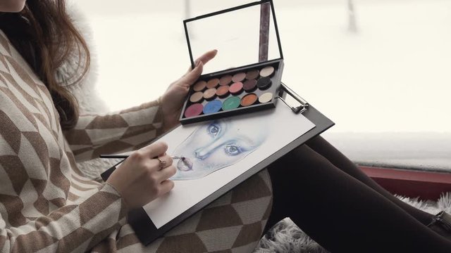 The artist on makeup and beauty sits on the window sill near window and creates a beautiful face makeup chart using watercolor paper, palette shadows, lipsticks and blushes. Face chart