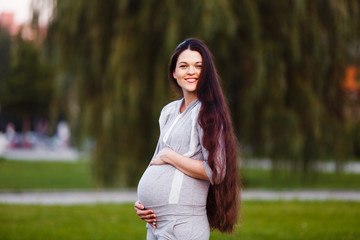 Young smiling pregnant woman in gray overalls posing in park. Holds on to his stomach.