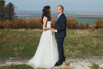 Wedding couple is hugging and holding hands in sunny day. Groom and bride in white satin lace dress are kissing with view on green and yellow field. Autumn nature panoramic landscape.