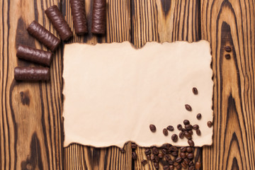 Row of yummy chocolate candies and fried halves of coffee beans near blank burnt paper on old weathered wooden table